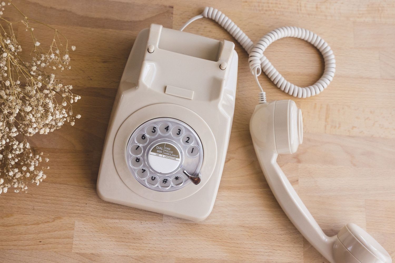 Vintage cream rotary dial telephone with handset off hook on wooden surface with dried white flowers.