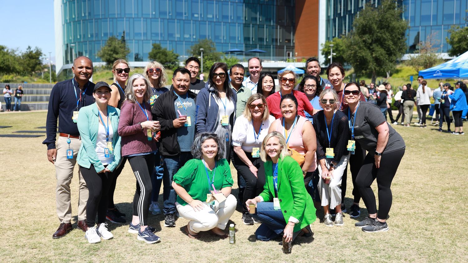 Large group of diverse professionals posing outdoors near a modern glass building on a sunny day.