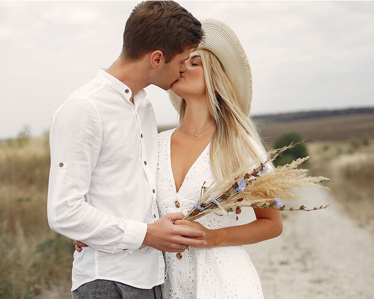 Romantic couple kissing outdoors, woman in white dress and hat holding wildflowers in a scenic field. Audio guestbook audiogram.