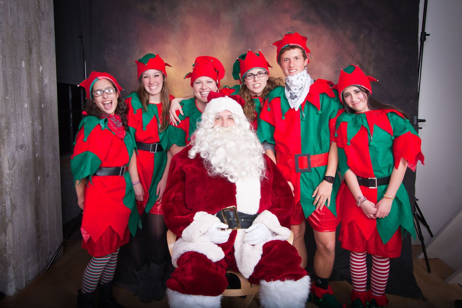 Santa Claus seated with six elves in red and green costumes posing for a festive Christmas photo.