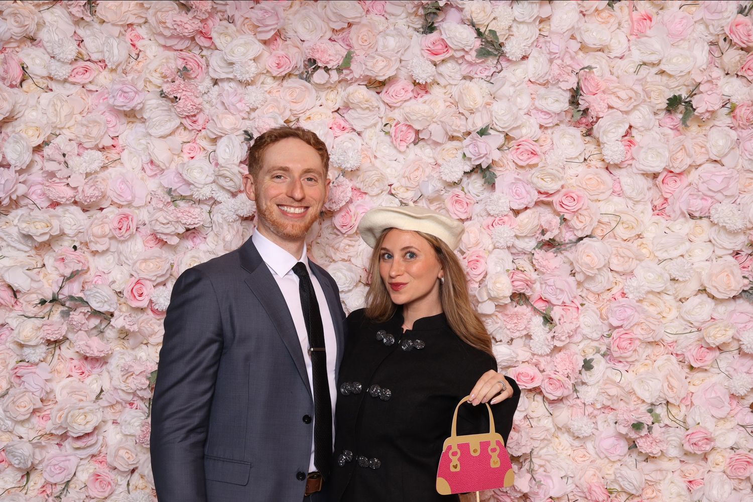 Couple posing in front of pink floral wall, woman in black outfit with white hat holding pink bag, man in gray suit.