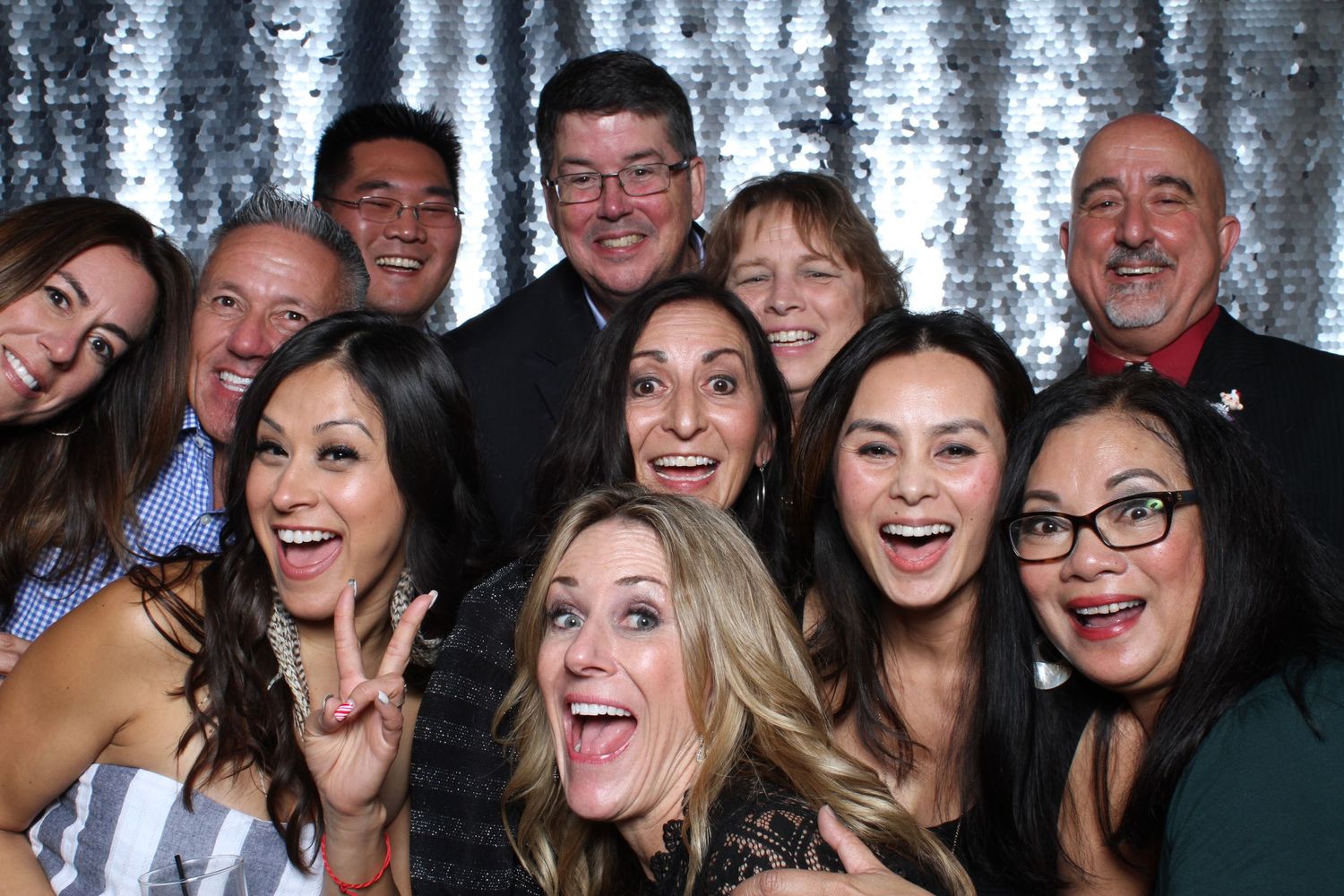 Group of smiling friends posing together in a photo booth with a silver sequin backdrop at a party.
