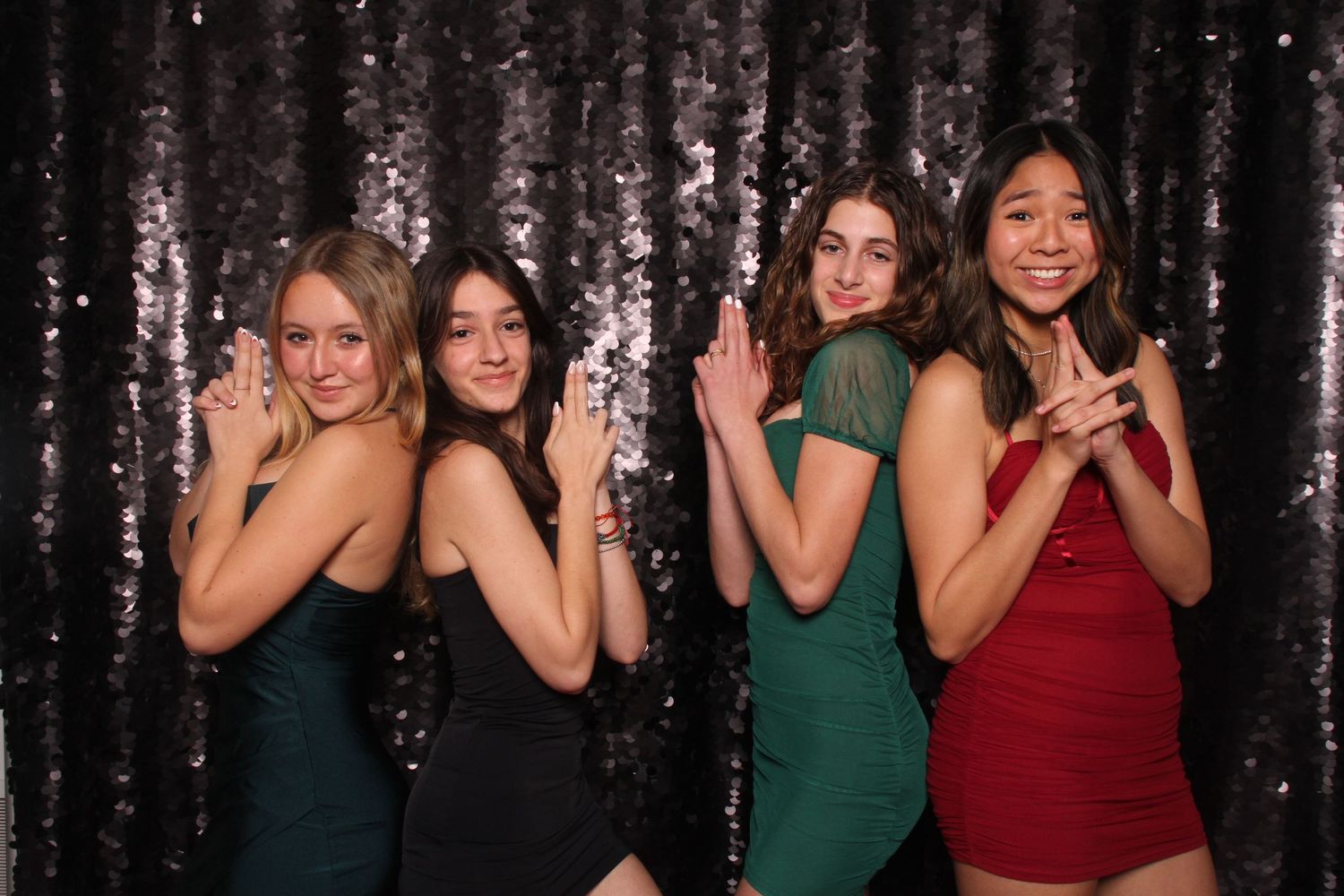 Four young women in formal dresses pose and smile in front of a black sequin backdrop during the Hillsdale High Prom at the Design Center.