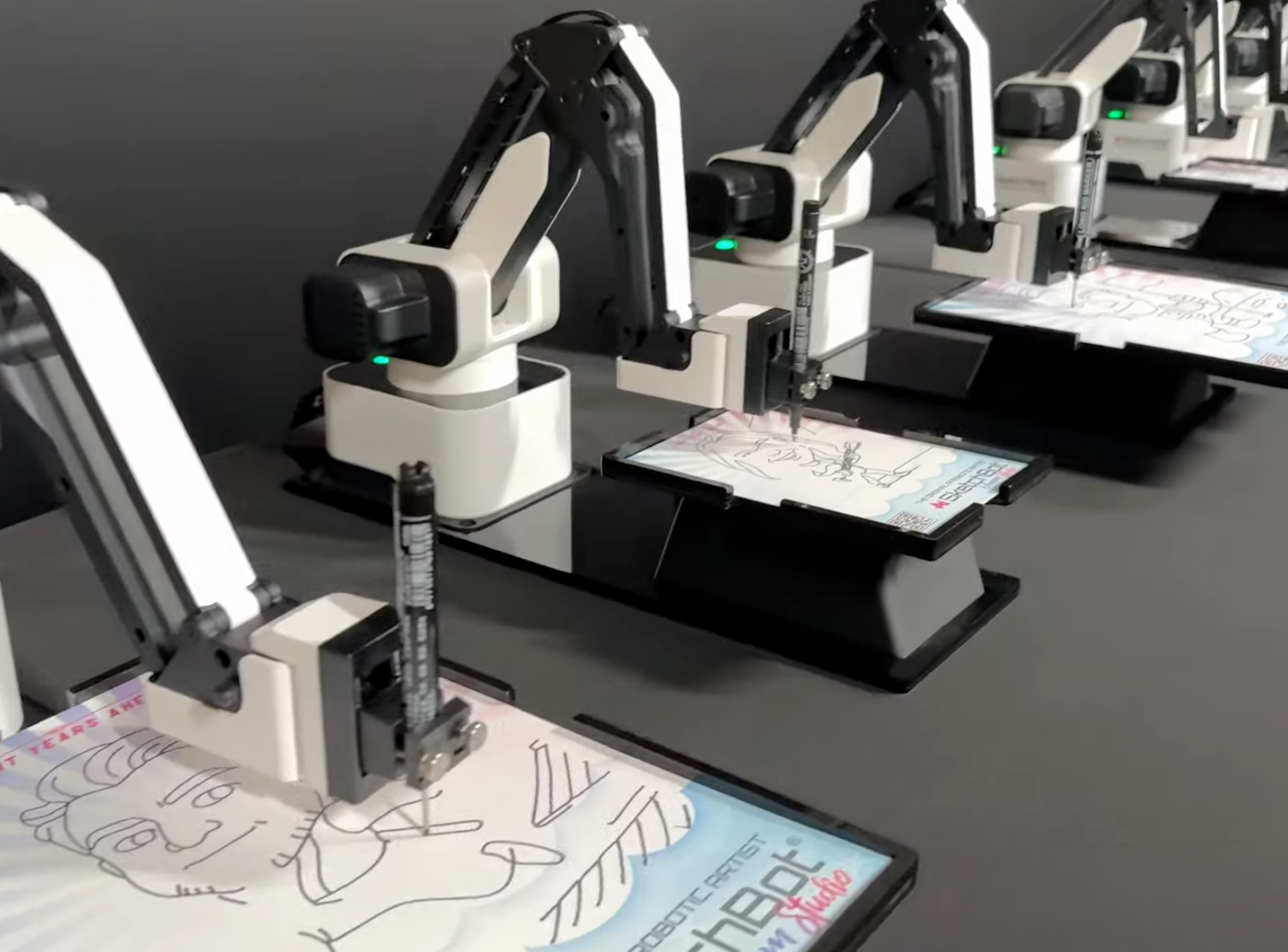 Robotic arms with drawing tools creating sketches on paper in an automated manufacturing demonstration.