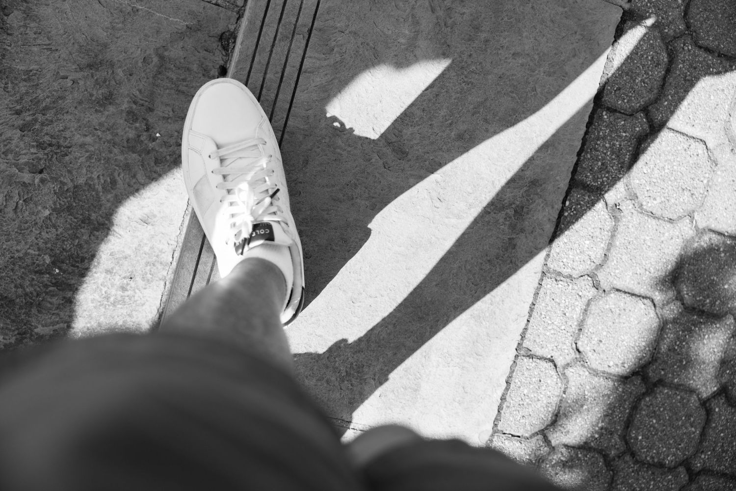 Black and white photo of a person wearing white sneakers standing on a cobblestone and pavement surface in sunlight.