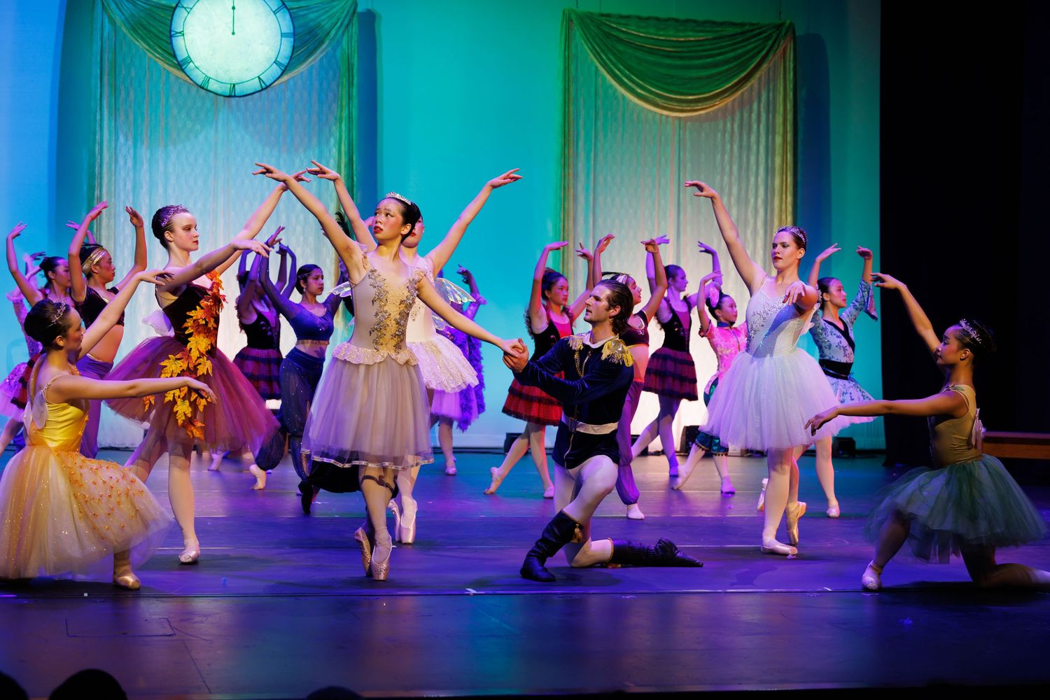 Ballet dancers perform on stage in colorful costumes during a theatrical production under a decorative clock backdrop.