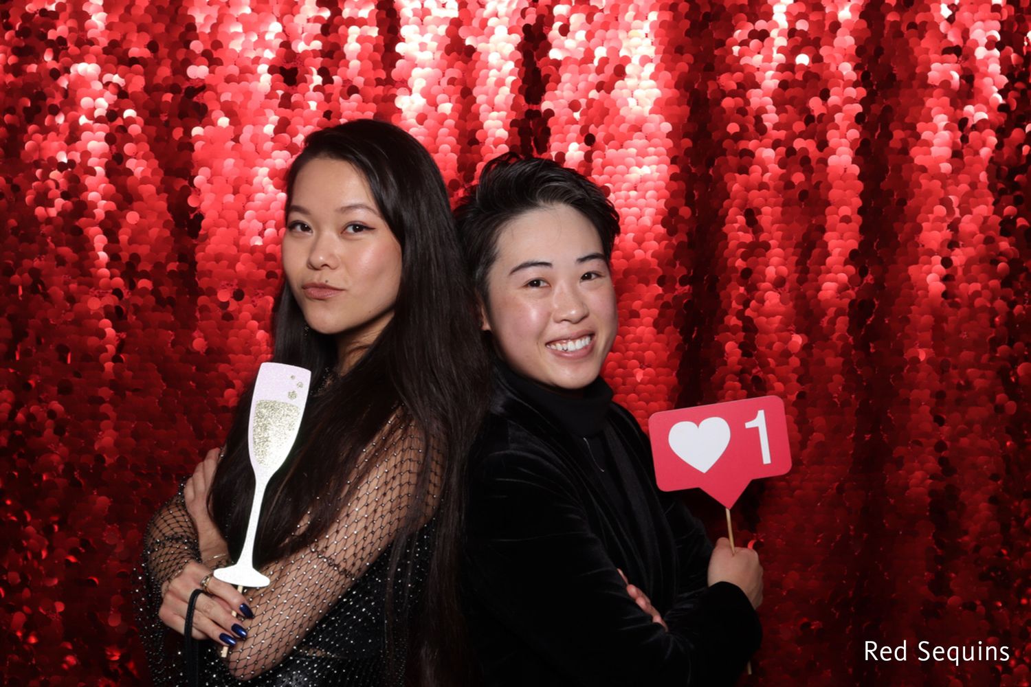 Two elegantly dressed women posing back-to-back holding champagne glass and heart props.