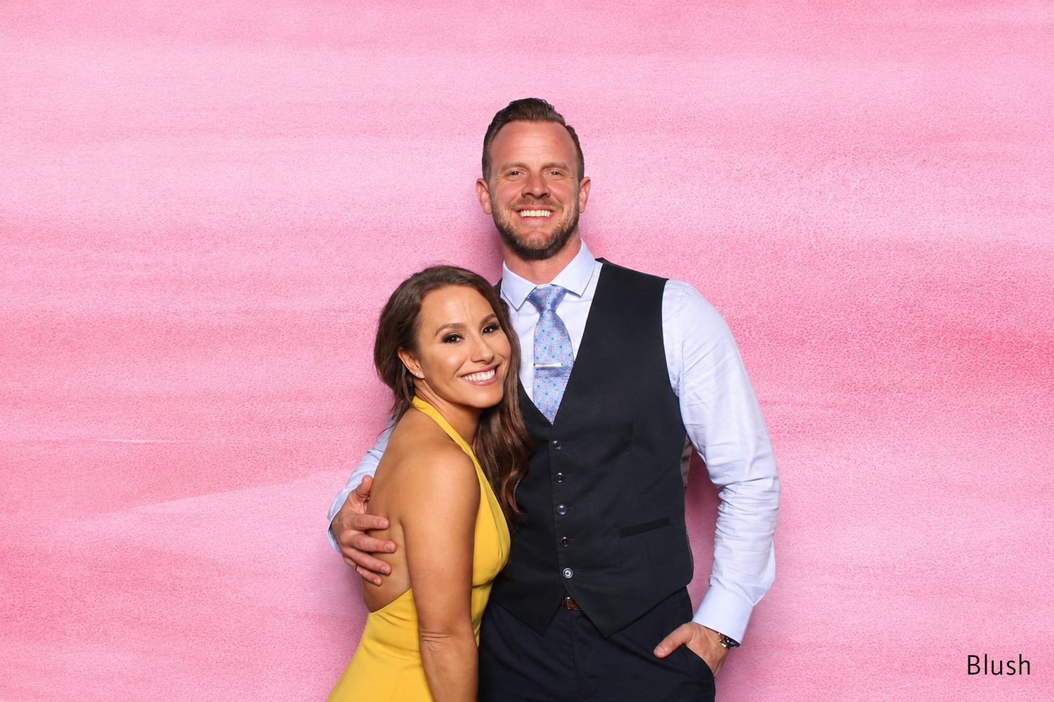 Smiling couple posing together in front of a pink blush photo booth backdrop.