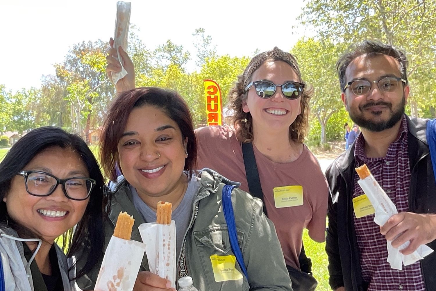 Four smiling friends holding snacks outdoors on a sunny day, wearing name tags at a casual outdoor event.