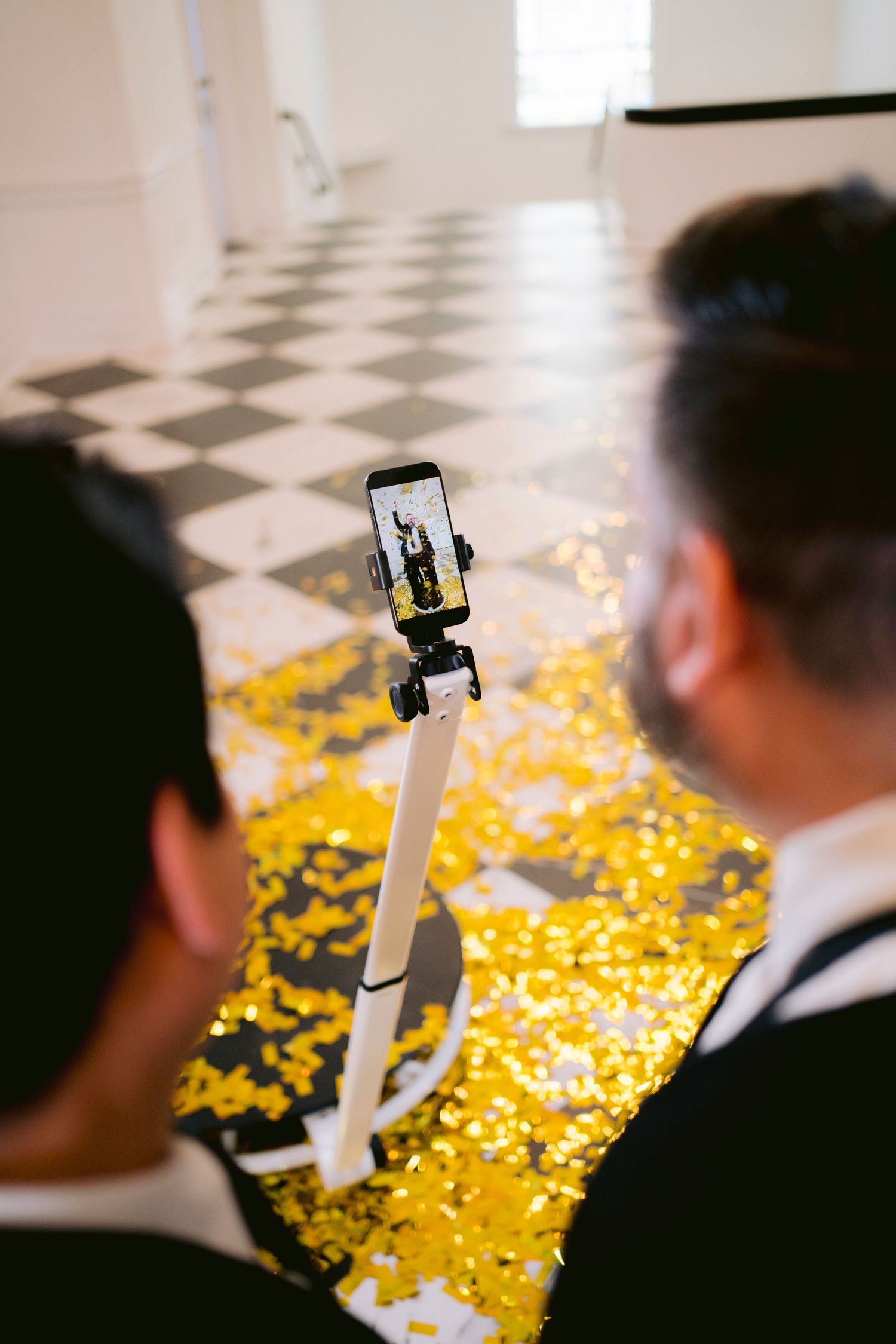 Two men in suits use a selfie stick to photograph golden confetti scattered on a black and white checkered floor.