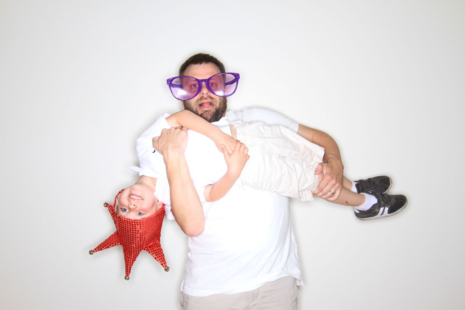 Man wearing oversized purple glasses holds a child upside down while both pose with a red star balloon prop.