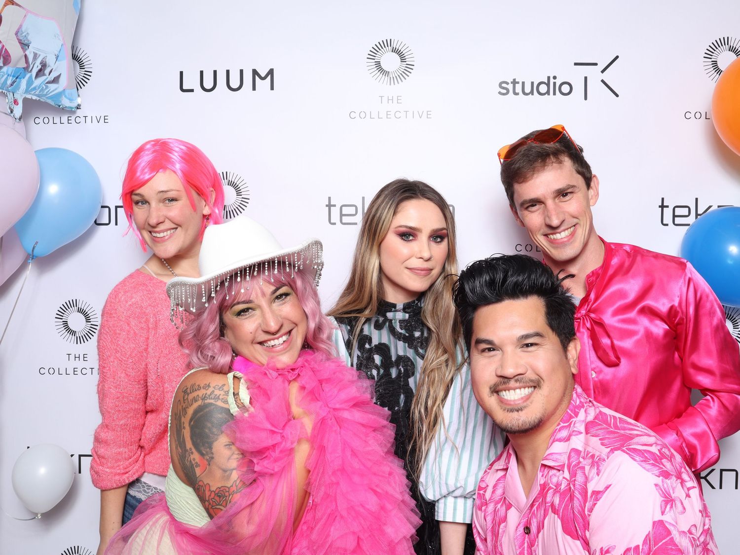 Five friends pose together at a colorful event with LUUM and Studio K banners, wearing vibrant pink outfits and accessories in San Francisco