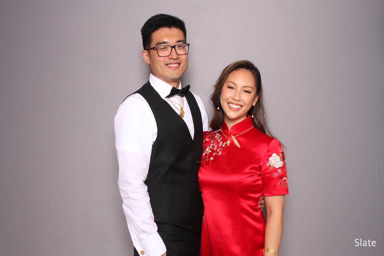 Couple posing in photo booth, man in vest and woman in red qipao against gray backdrop.