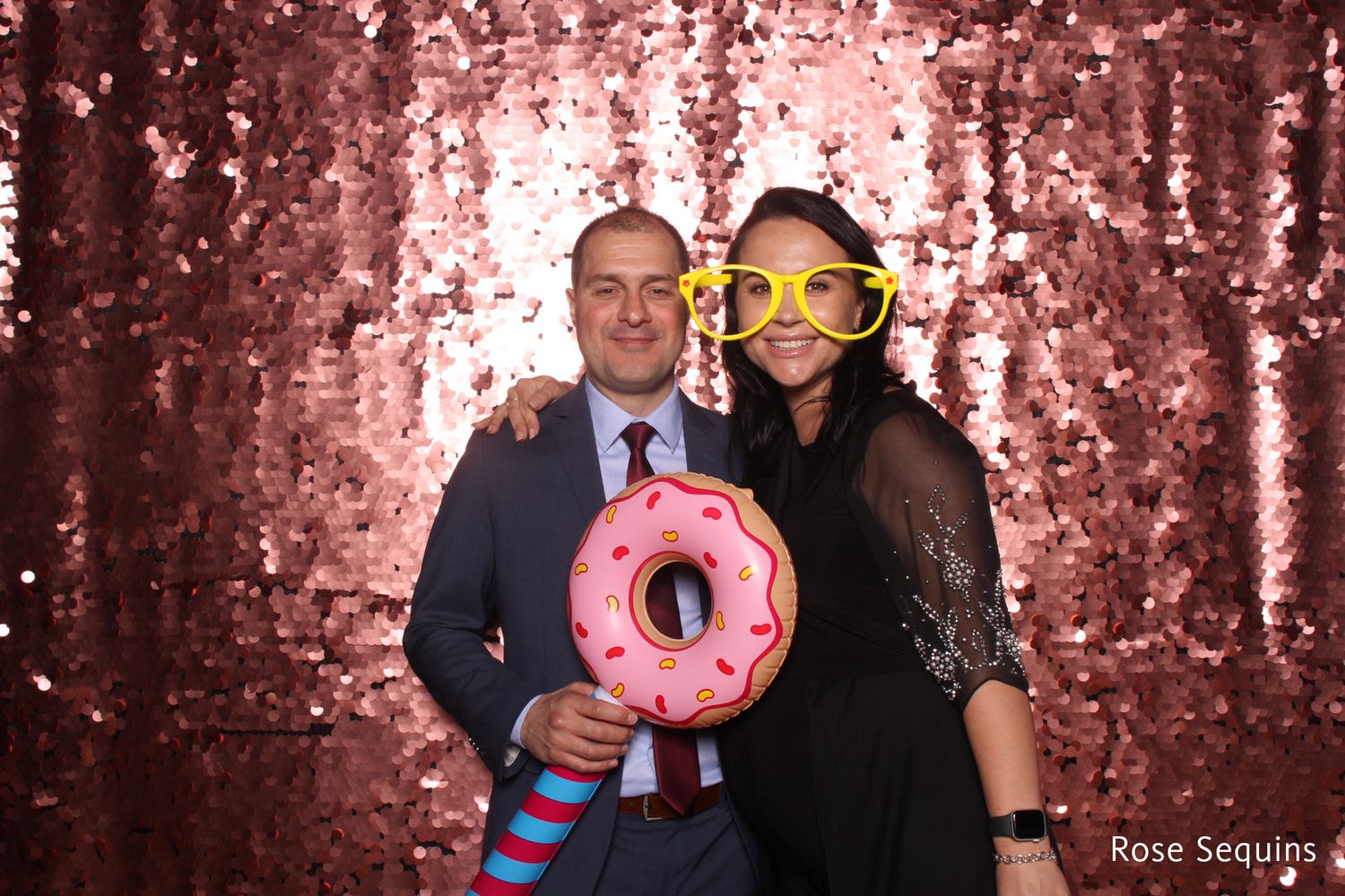Couple posing with a giant donut prop and star-shaped glasses in front of a rose sequins backdrop.