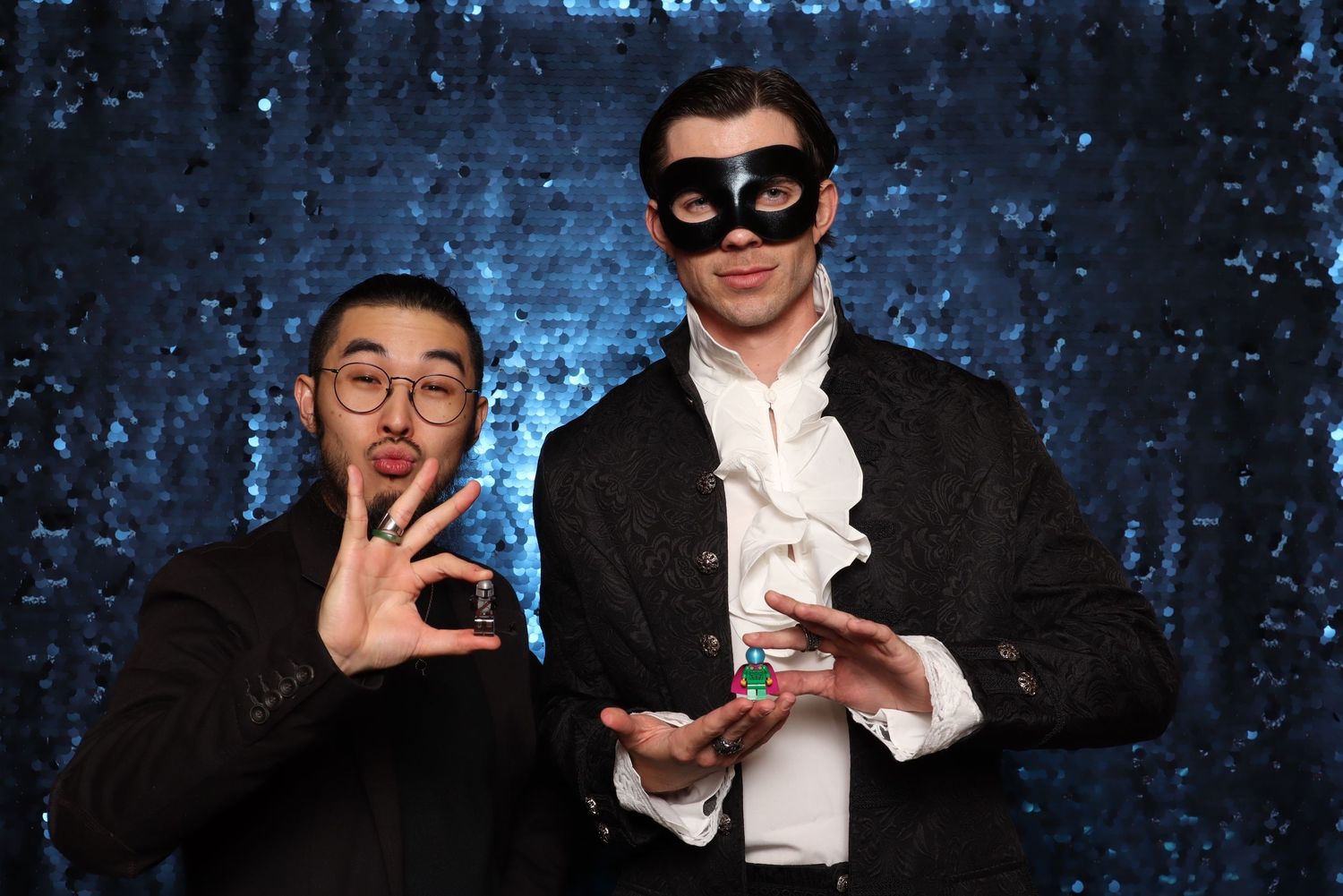 Two men posing at a masquerade party, one wearing a black eye mask and tuxedo, against a blue sequin backdrop. Vagaro Party at Ruby Hill