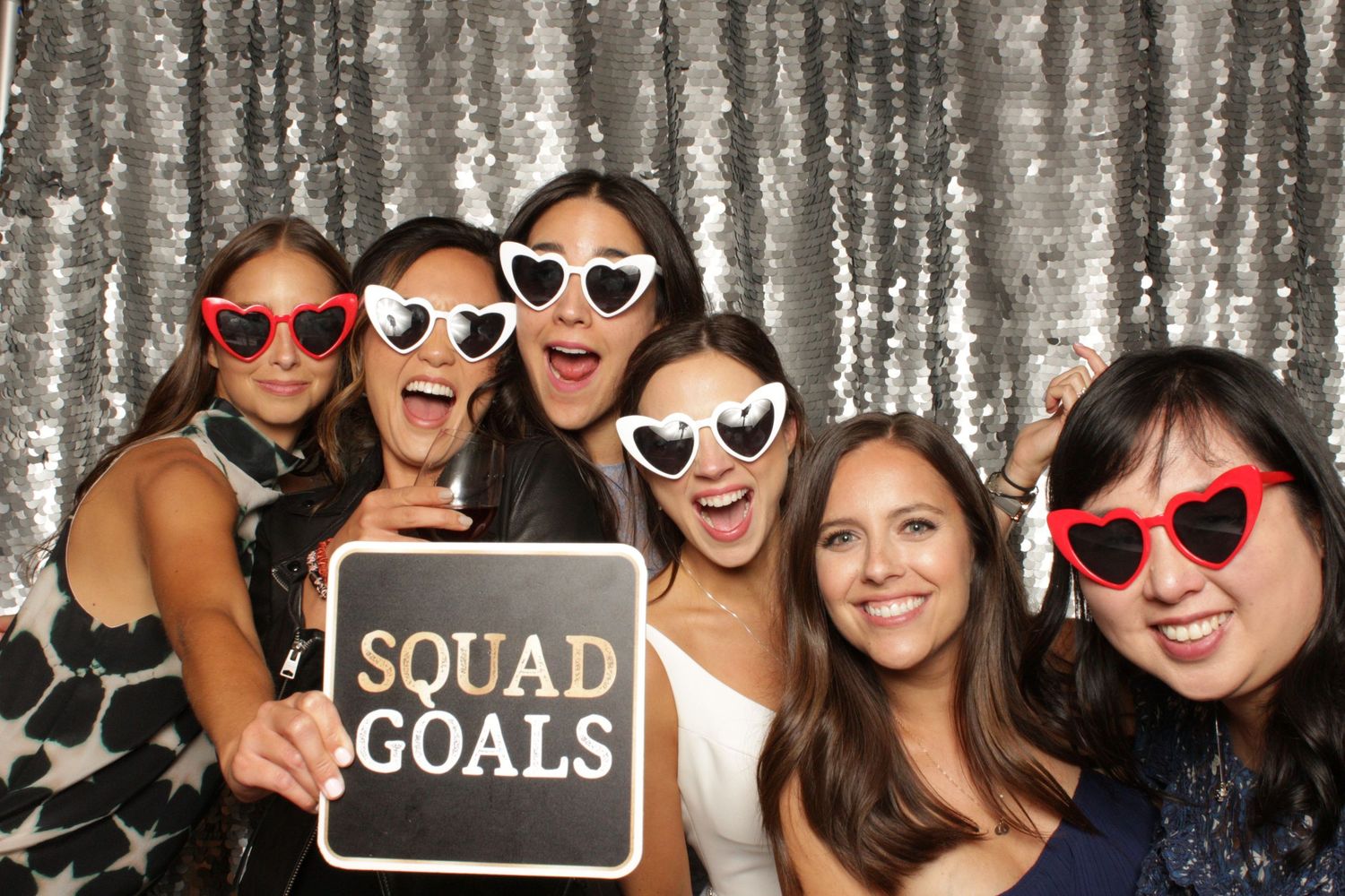 Women posing in a photo booth with heart-shaped sunglasses and a Squad Goals sign against a silver sequin backdrop.