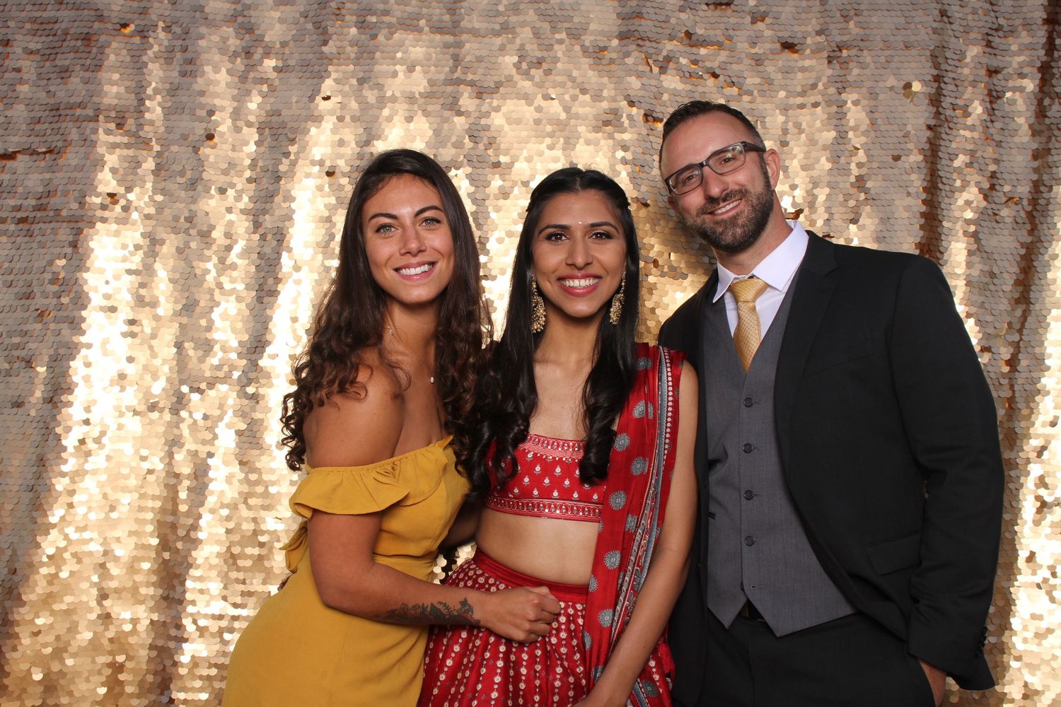 Three guests pose at a wedding photo booth with gold sequin backdrop.