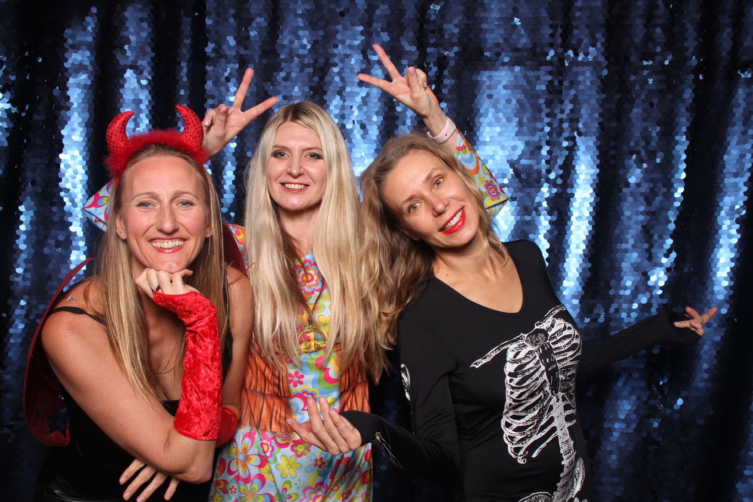 Three women in Halloween costumes pose playfully in front of a blue sequin backdrop at a festive party.