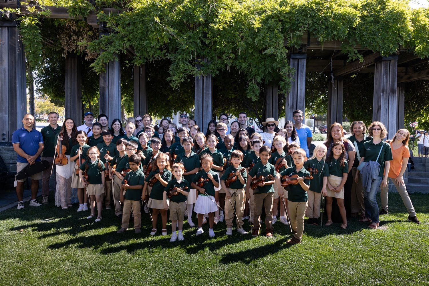 Large group of children in green uniforms and adults posing outdoors near vine-covered columns on a sunny day.