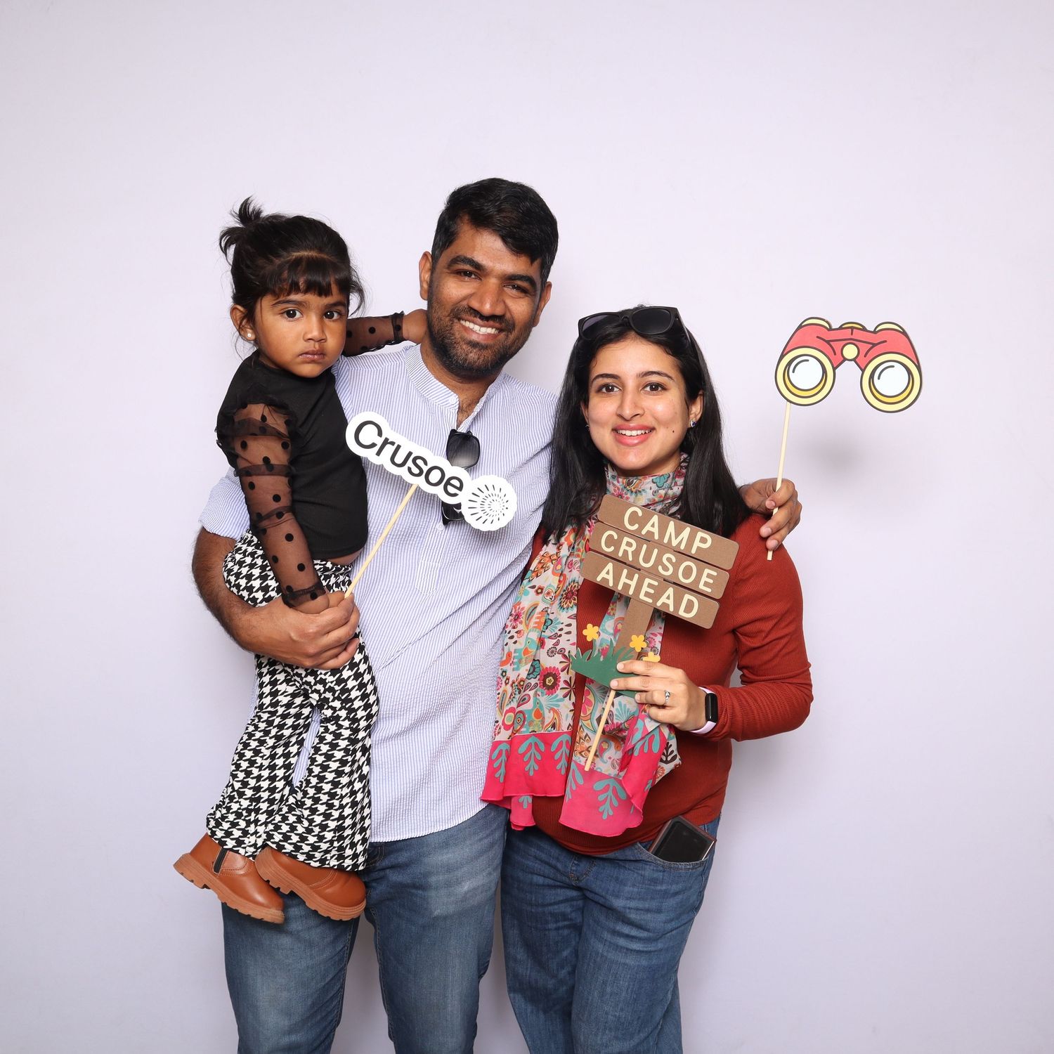 Family of three posing with Camp Crusoe props, parents holding signs and binoculars cutout, child in checkered dress.