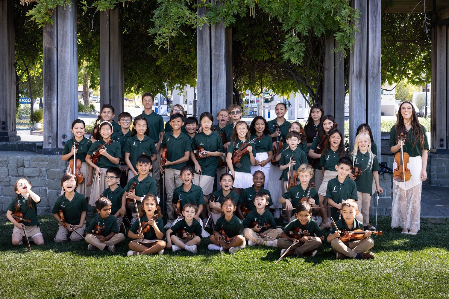 Young students in green uniforms pose outdoors with violins and string instruments for a school orchestra group photo.