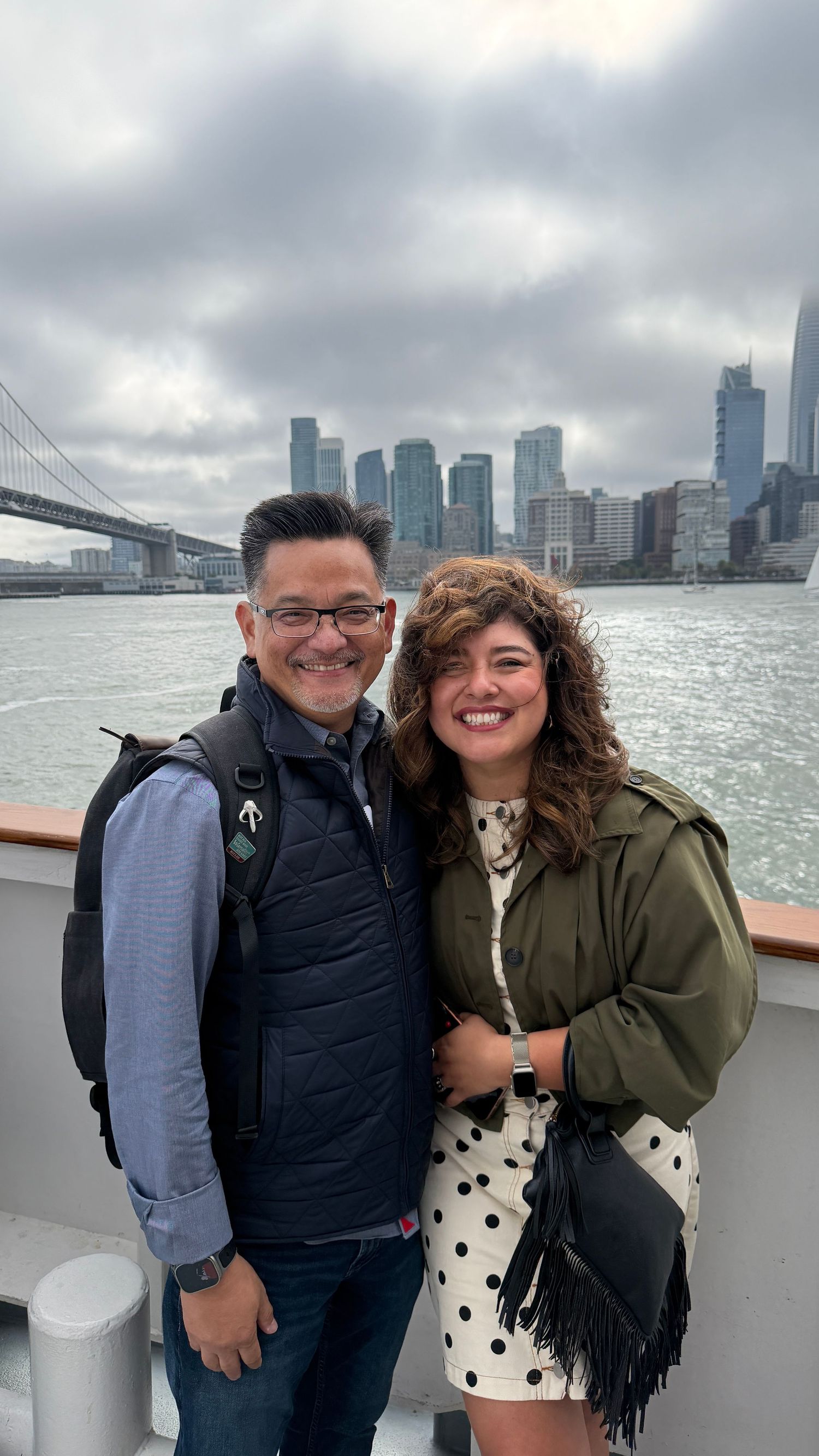 Couple smiling on a ferry with NYC skyline and Brooklyn Bridge in the background on a cloudy day.