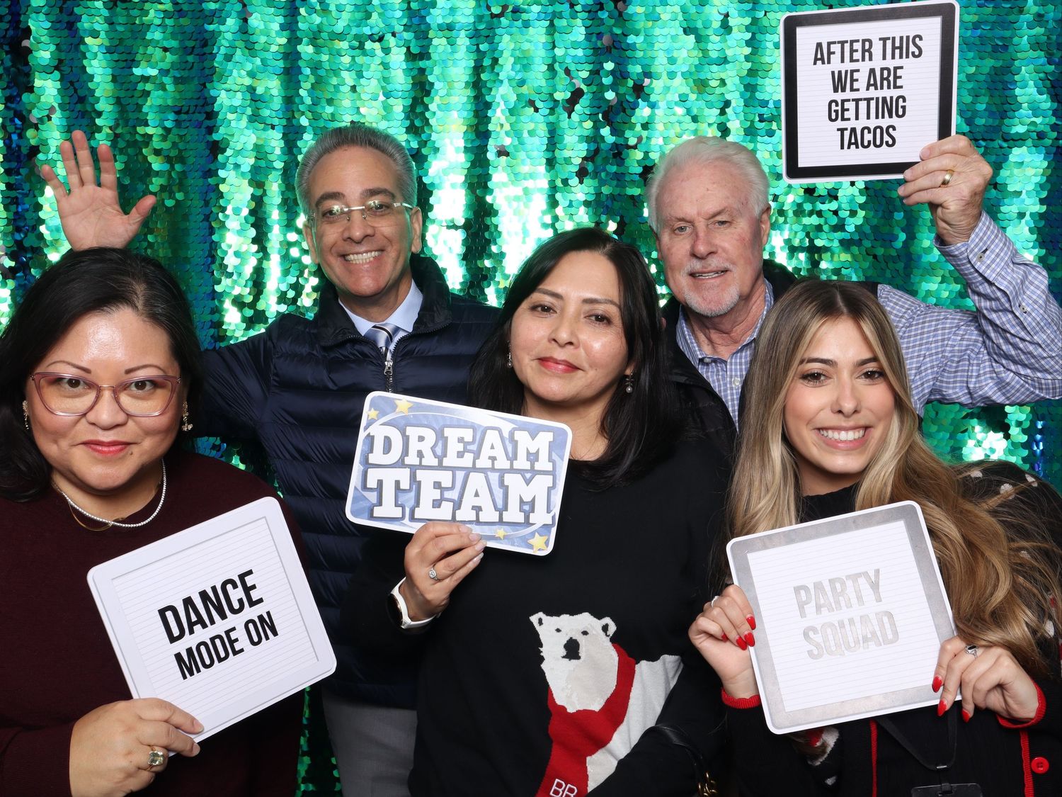 Five friends pose at a photo booth with fun signs including Dream Team, Dance Mode On, and After This We Are Getting Tacos. Gensler Holiday