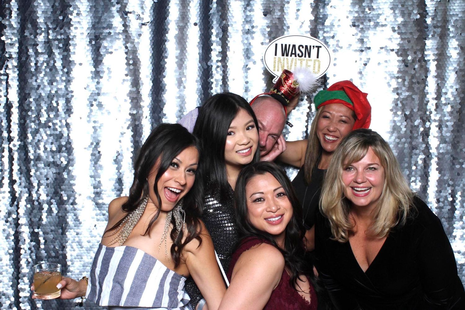 Group of six friends posing and smiling at a festive photo booth with silver sequin backdrop.