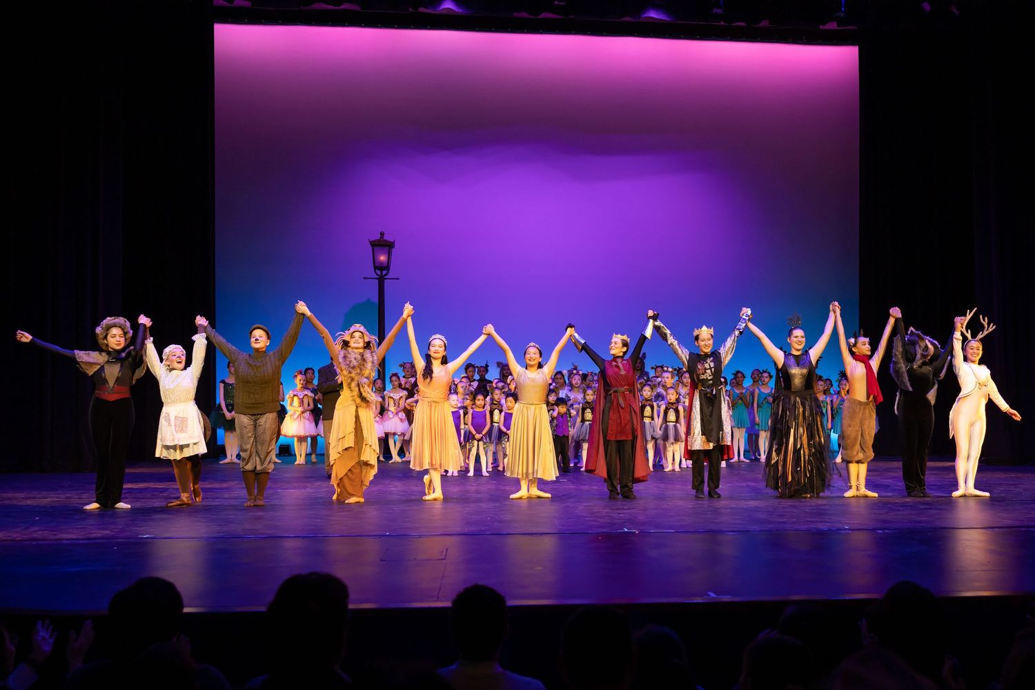 Theater cast takes a bow on stage, holding hands under purple lights in colorful costumes during a finale curtain call. Las Positas College