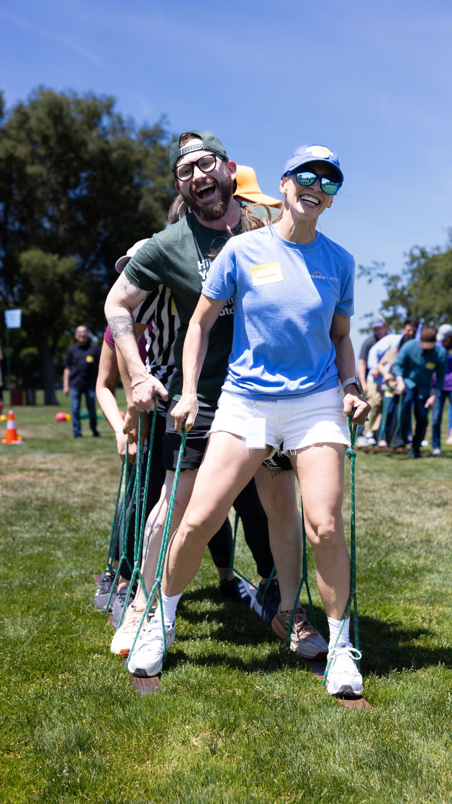 People laughing and competing in a three-legged race on grass at an outdoor team-building event on a sunny day.