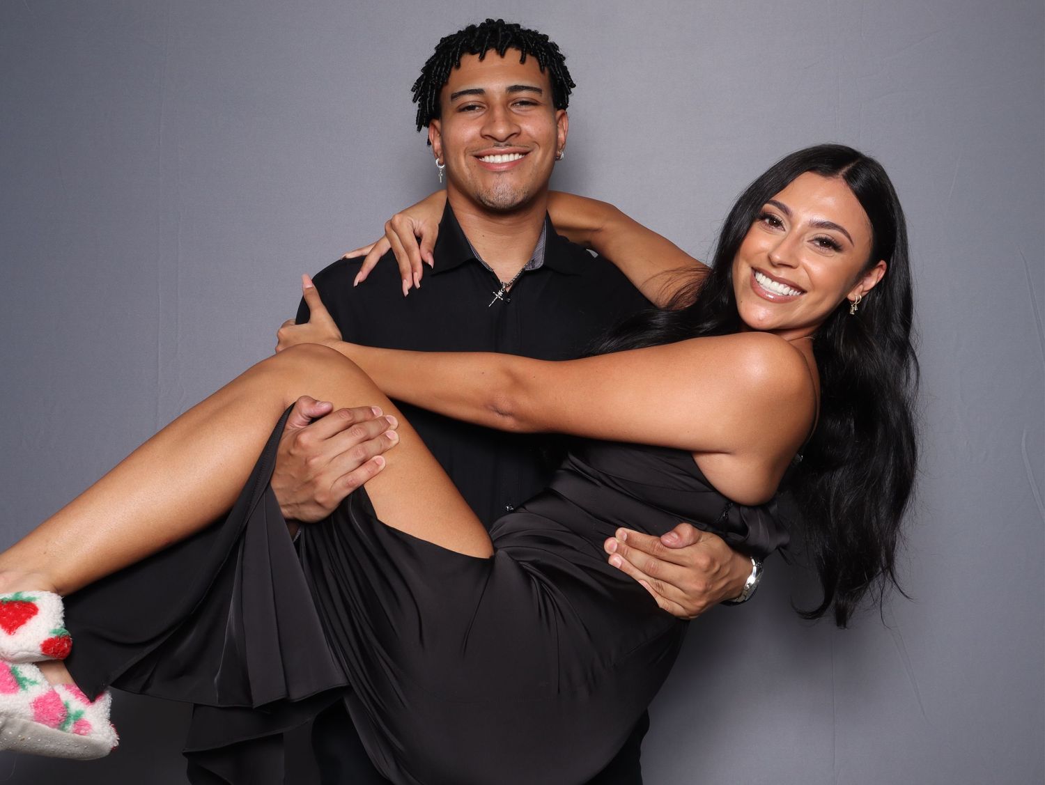 A smiling couple poses together in matching black outfits, the man holding the woman in his arms against a gray background.