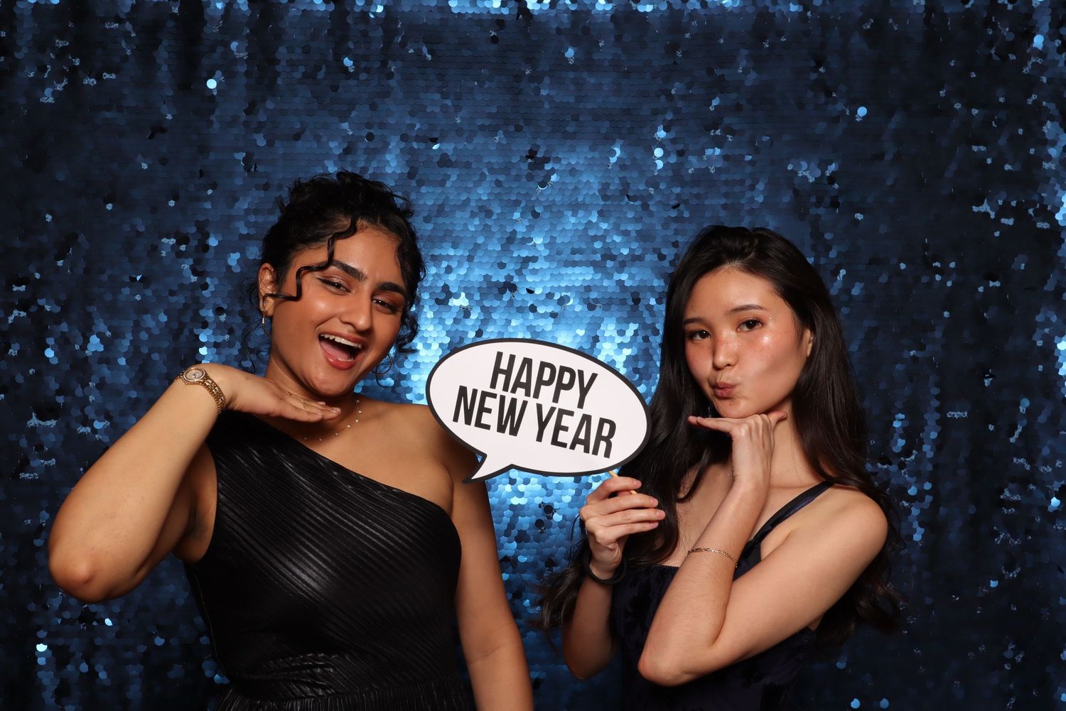 Vagaro Party at Ruby Hill. Two women pose at a New Year's Eve photo booth with a Happy New Year sign against a blue sequin backdrop.