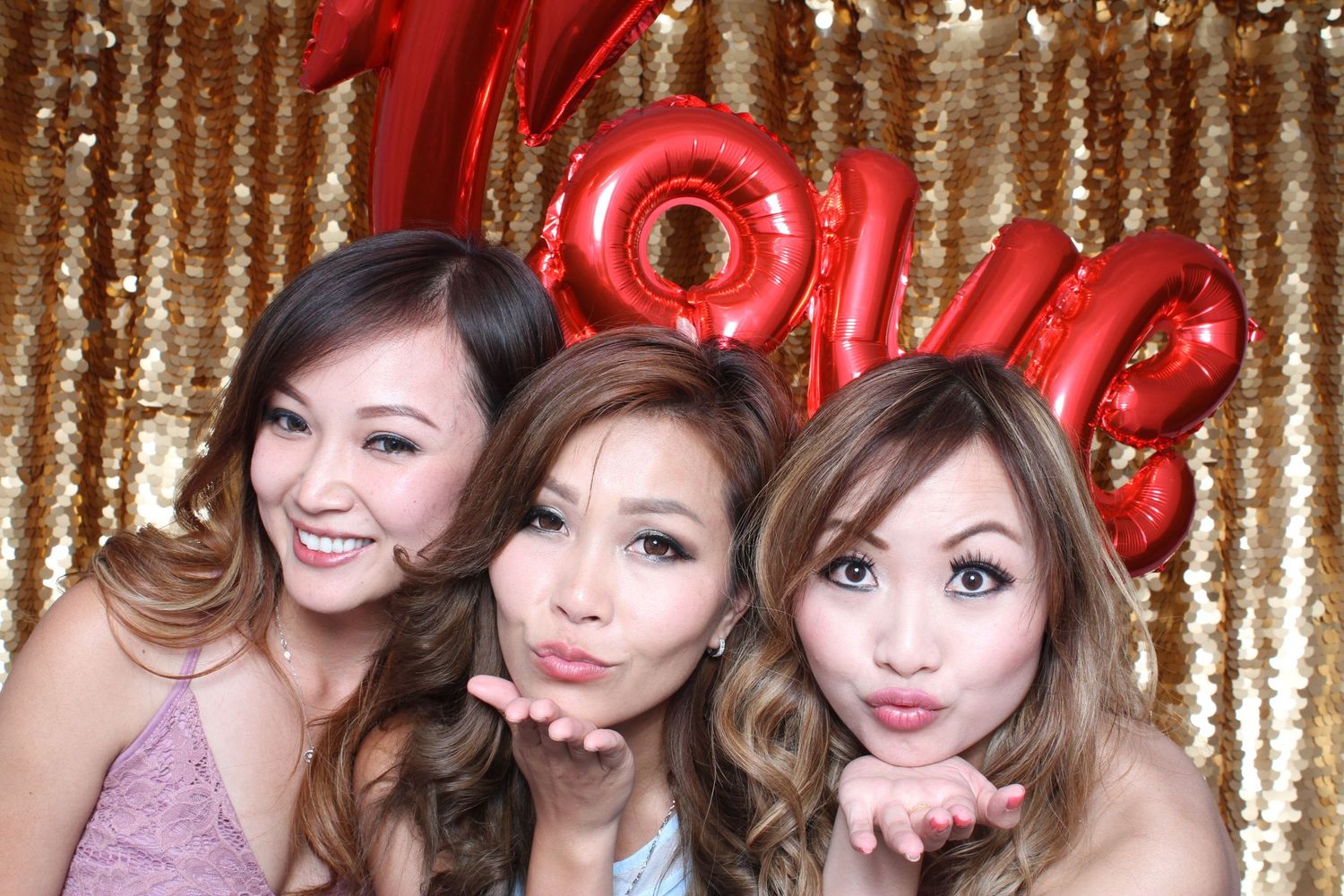 Three young women pose together blowing kisses in front of a gold sequin backdrop with red love balloons.