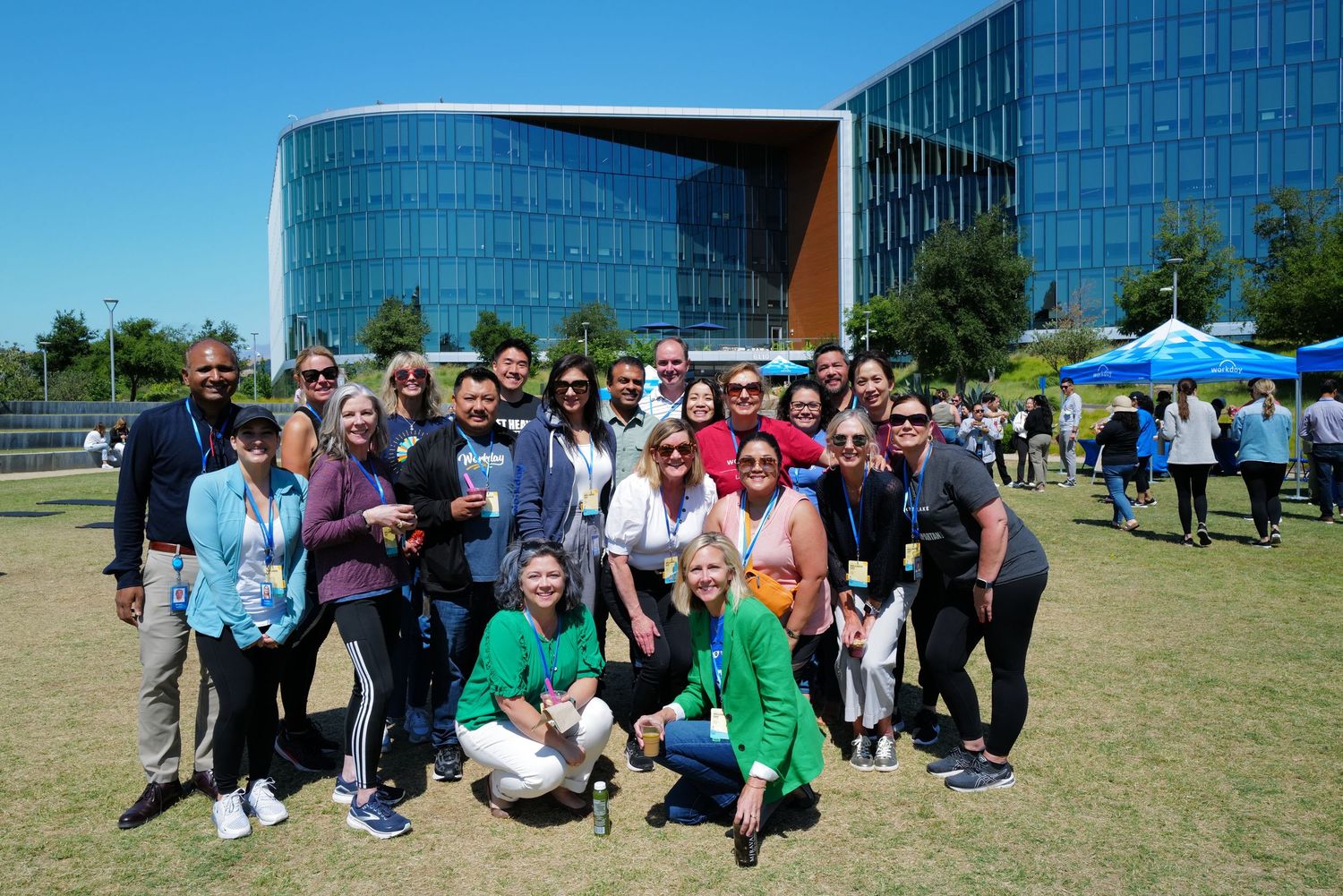 Large group of smiling professionals posing outdoors near a modern glass building on a sunny day.