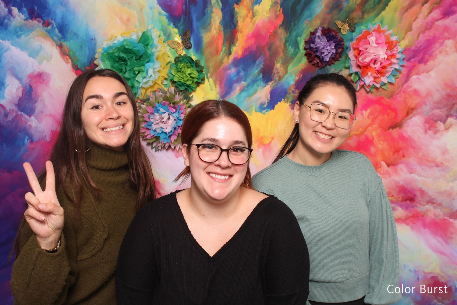 Three young women smiling in front of colorful floral burst photo booth backdrop.