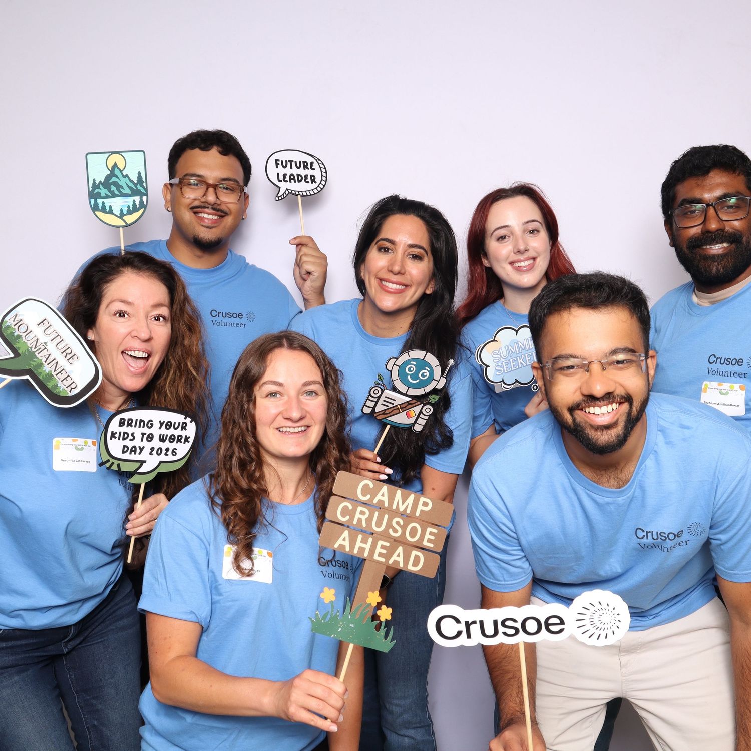 Group of seven Crusoe Energy employees smiling in blue t-shirts holding fun photo booth props at a Camp Crusoe event.