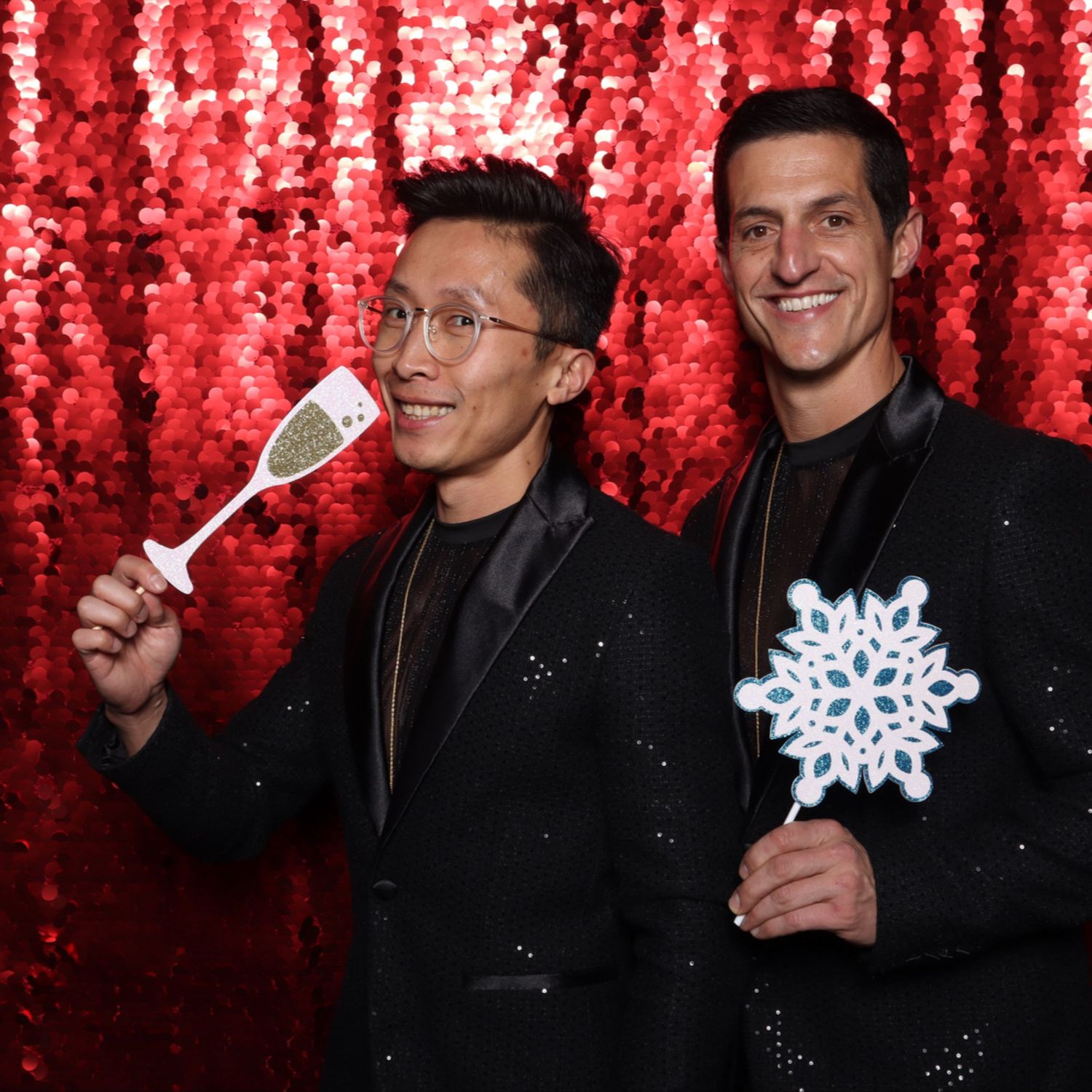 Two men in black sequin blazers pose with champagne and snowflake props in front of a red sequin backdrop. Gensler Holiday Party Fillmore