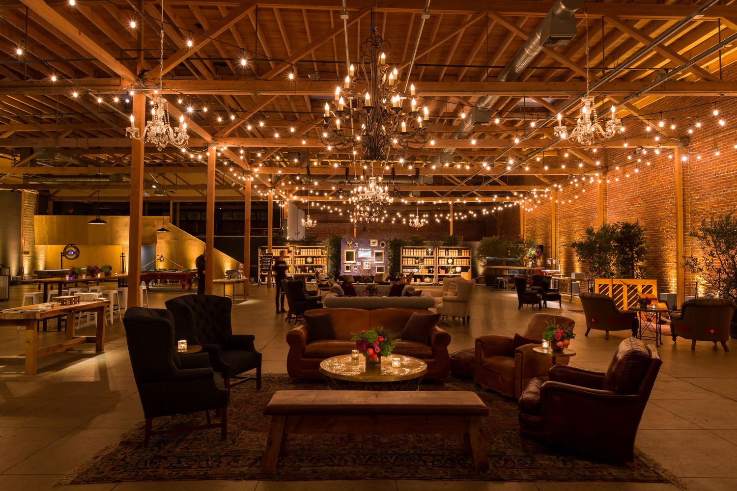 Elegant event venue with string lights, chandeliers, cozy seating, and warm amber lighting in a rustic industrial space.