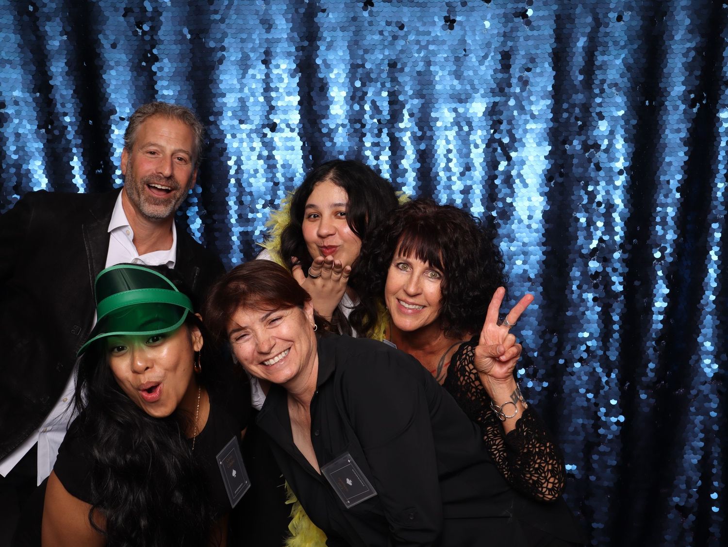 Group of five friends posing and making fun gestures in front of a blue sequin backdrop at a festive event.