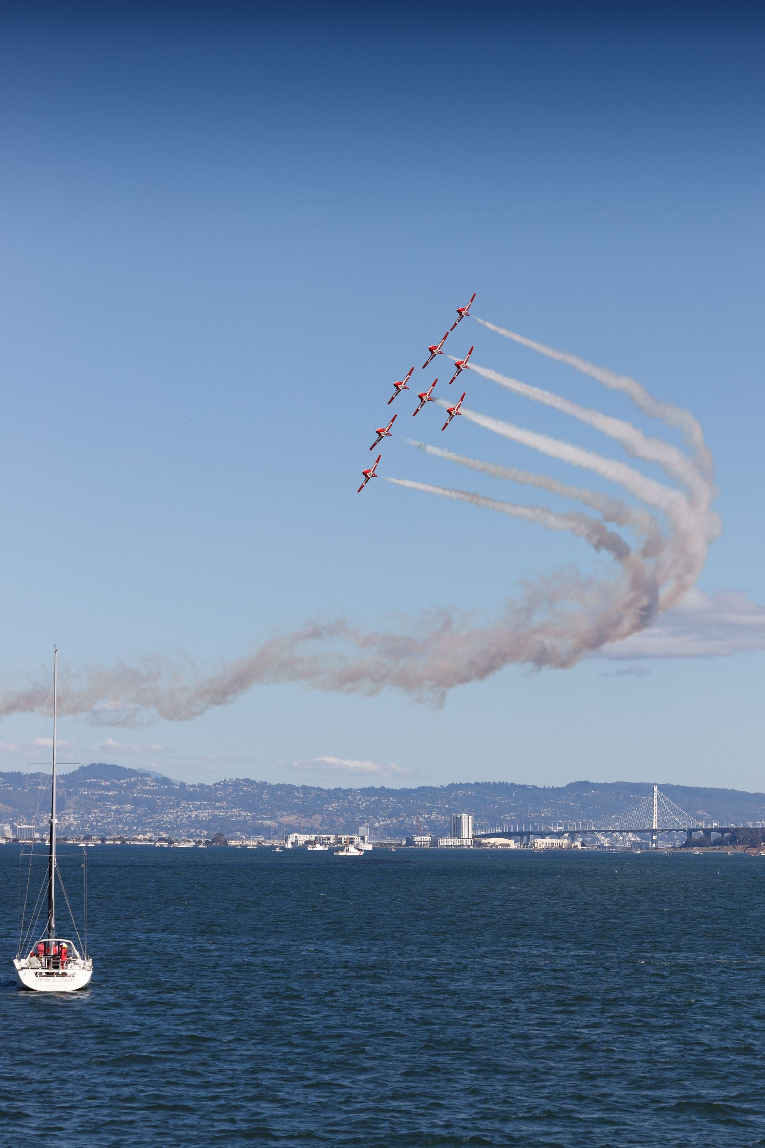 Formation of jets performing aerobatic display over San Francisco Bay, trailing white smoke, with sailboat and city skyline below.