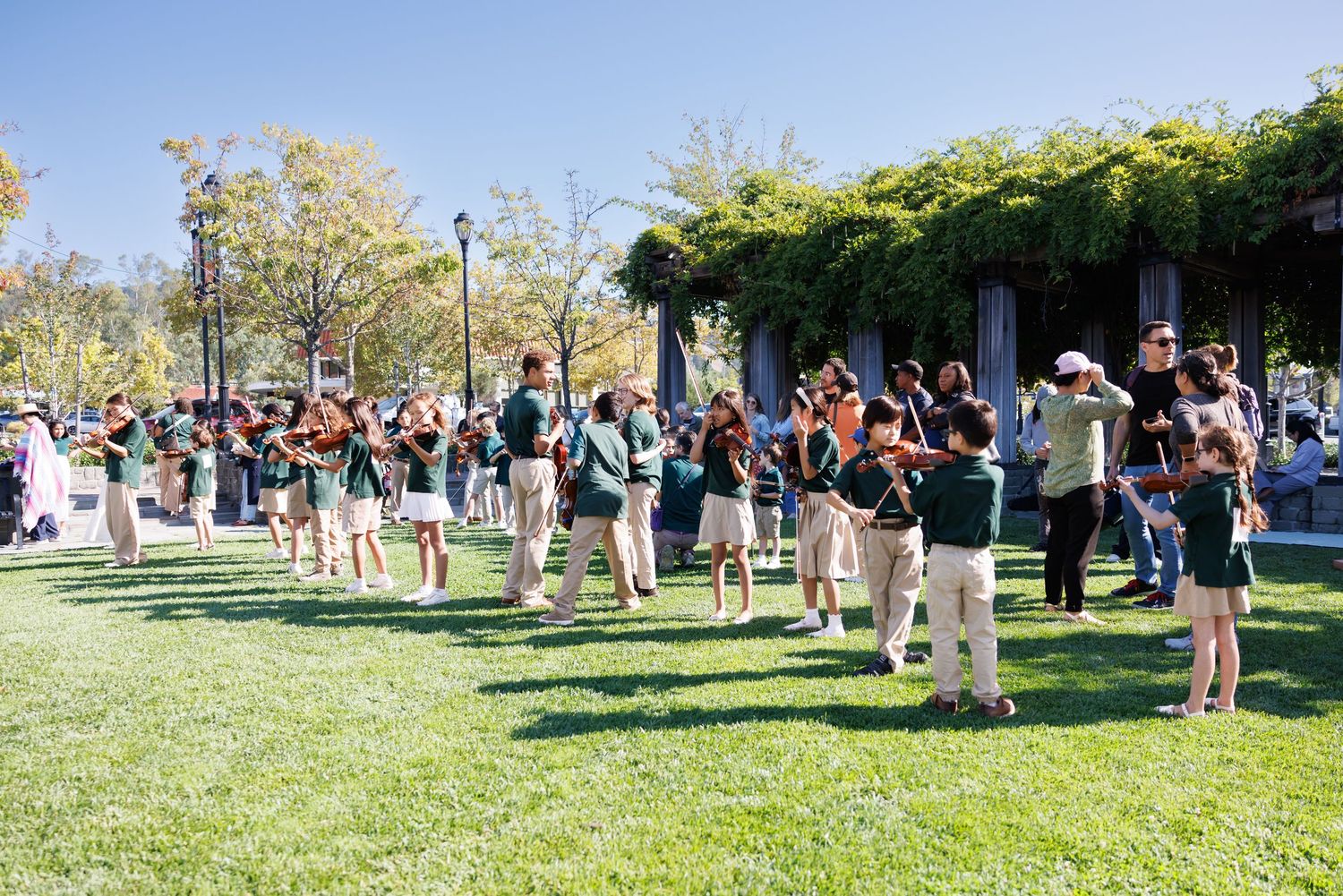 Studio Violin in Lafayette. Students in green uniforms play instruments on a sunny green lawn near a vine-covered pergola outdoors.