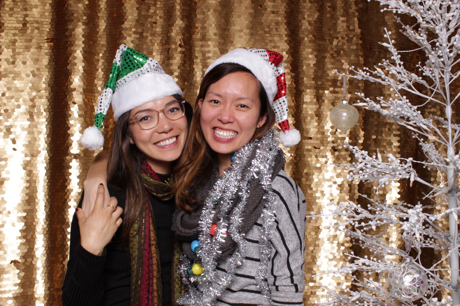 Two women wearing festive Christmas Santa hats smile together near a silver holiday tree with gold sequin backdrop.