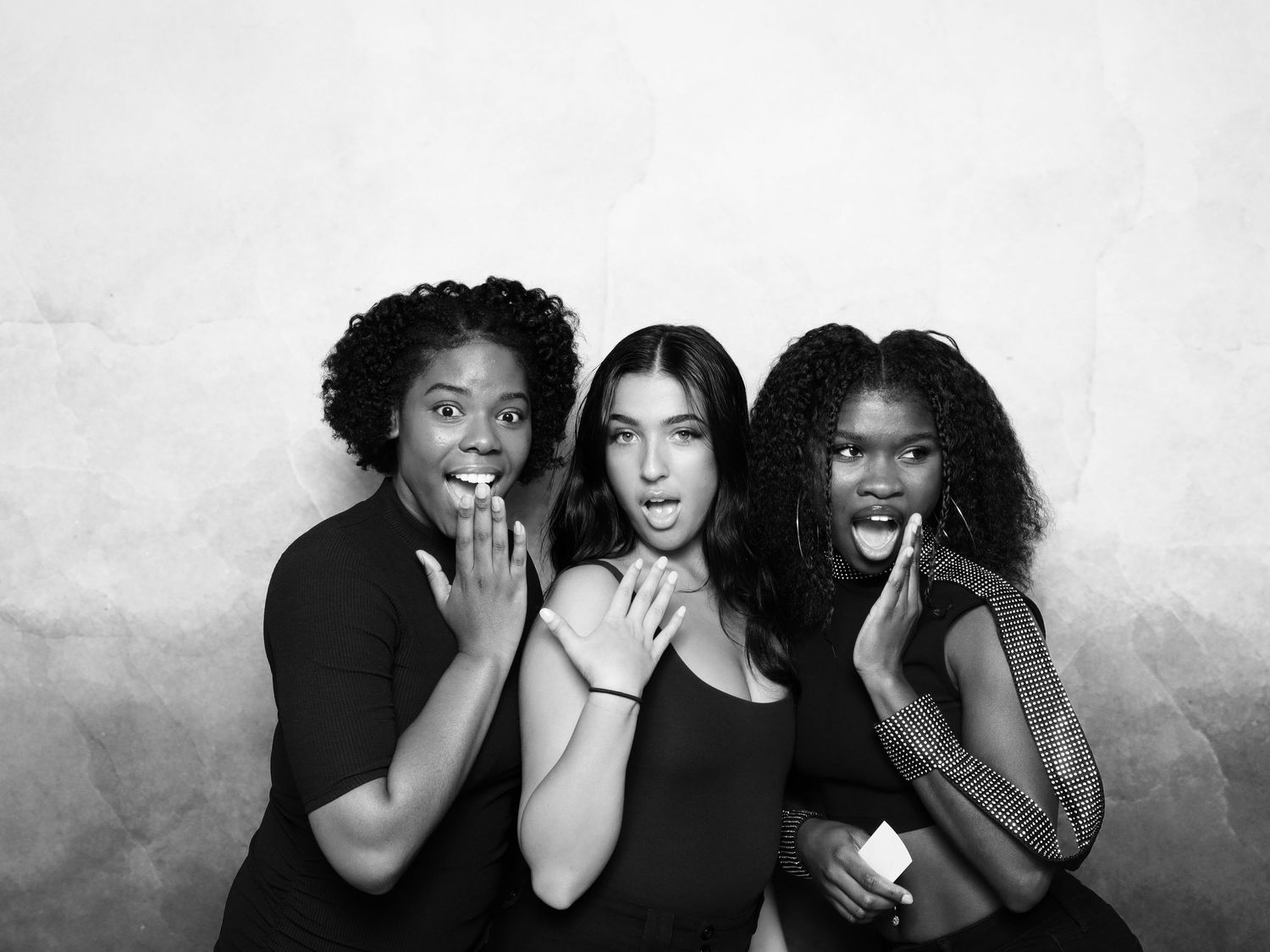 Three young women in black outfits pose playfully in a black and white photo booth portrait.