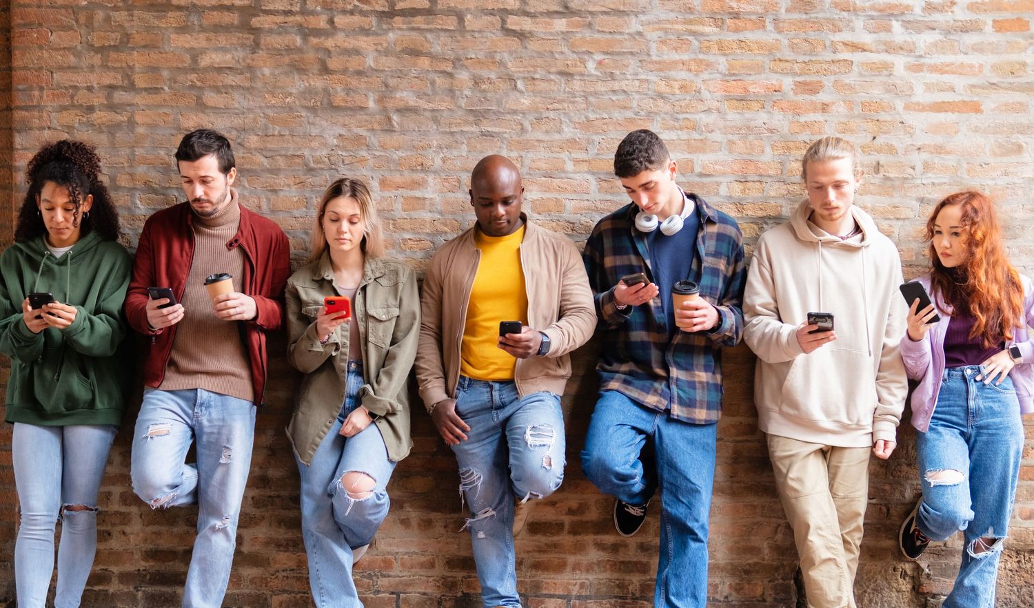 Diverse group of young adults leaning against brick wall, all looking at smartphones, representing social media addiction.