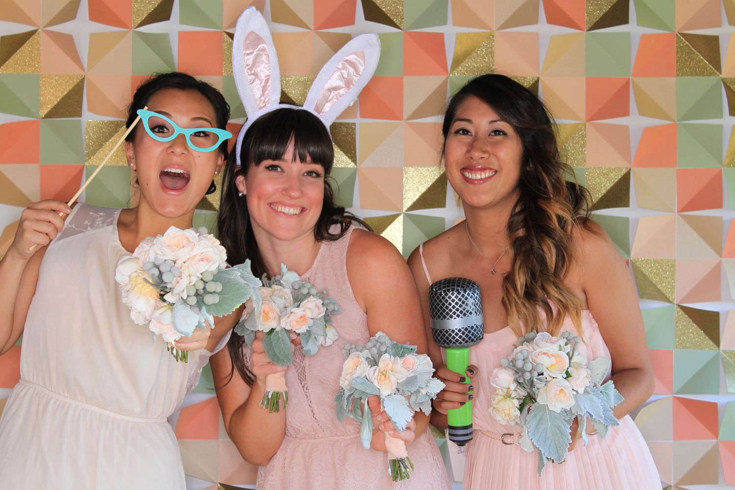Three bridesmaids in pink dresses pose with bunny ears, glasses props, and bouquets in front of a geometric backdrop.