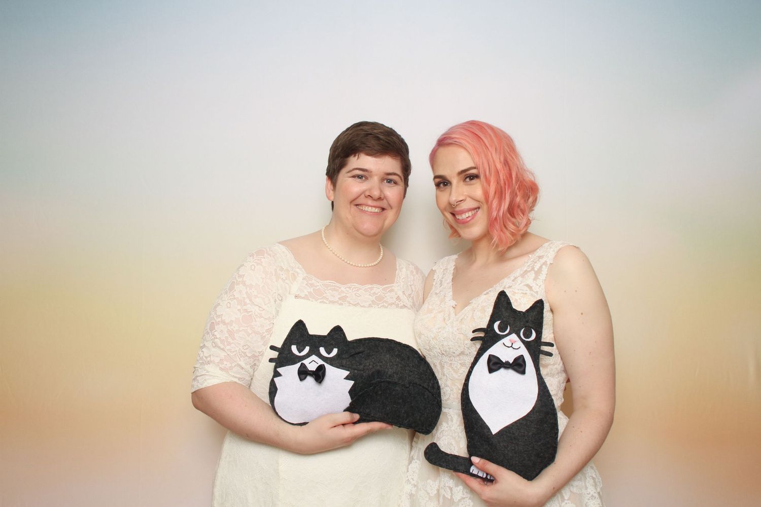 Two women in white dresses holding black and white cat plushies posing together against a pastel rainbow background.