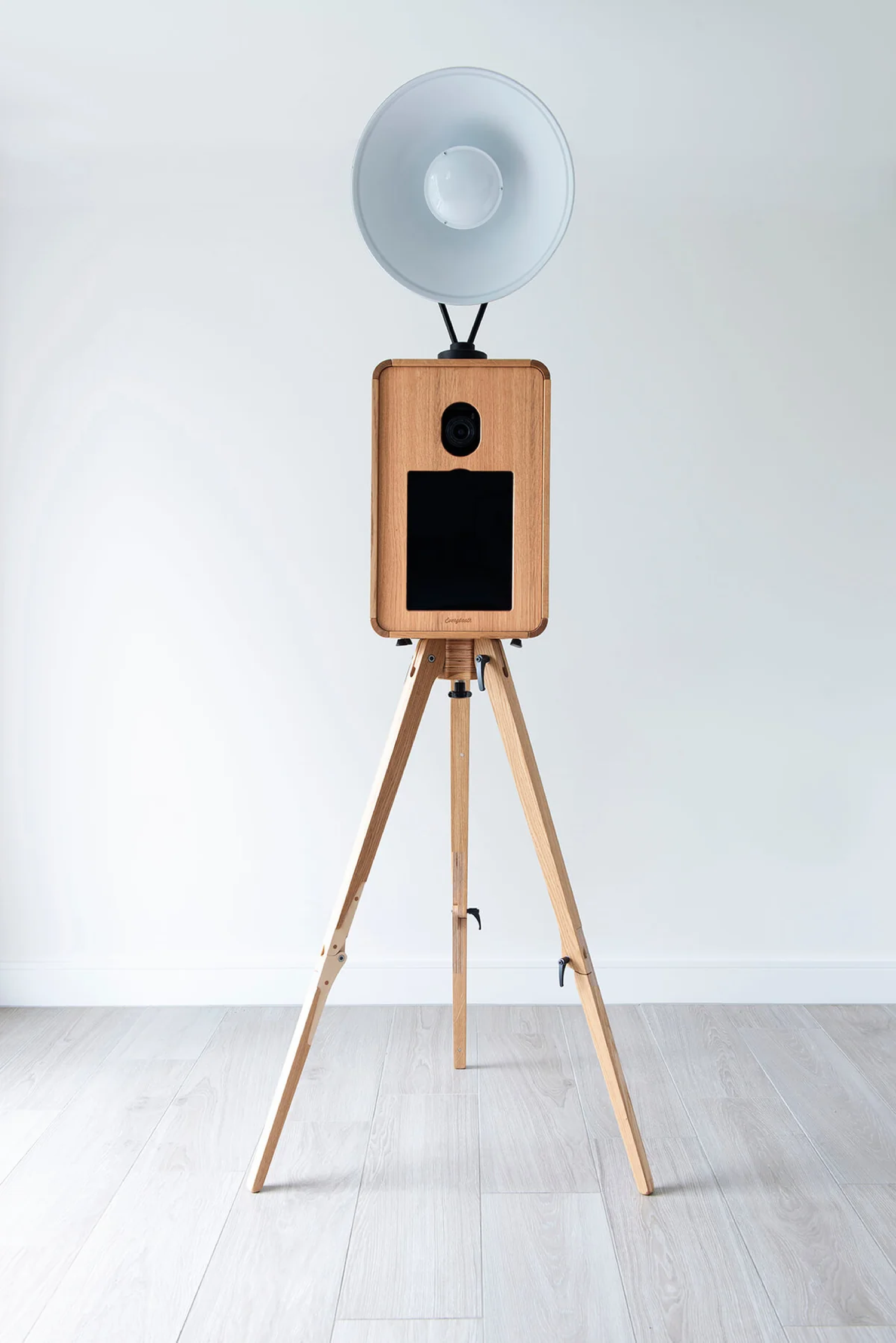 Wooden photo booth on a tripod with a large flash light on top, set against a white wall on a light wood floor. Ancienne booth