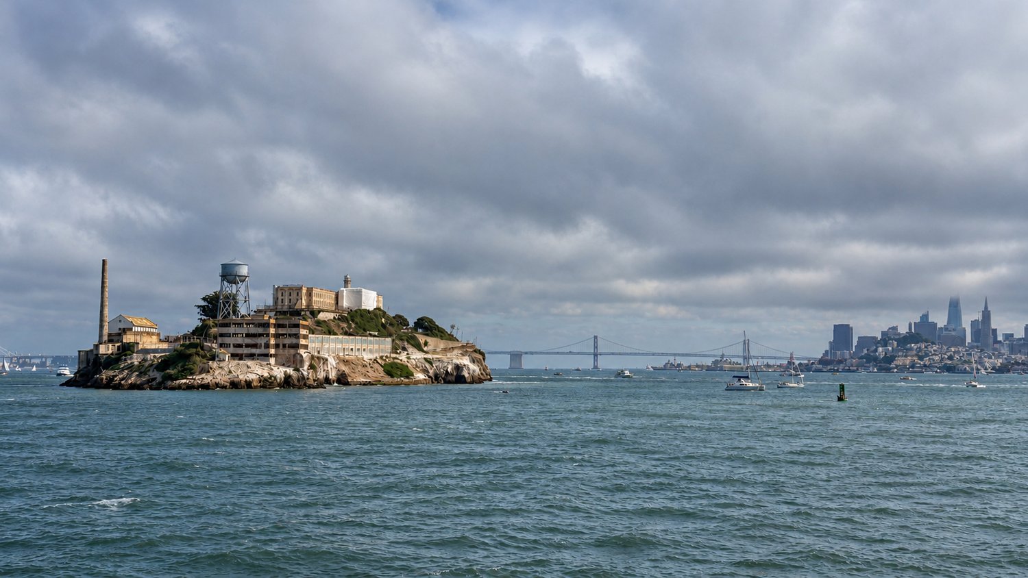 Aerial view of Alcatraz Island in San Francisco Bay with the city skyline and Bay Bridge visible under cloudy skies.