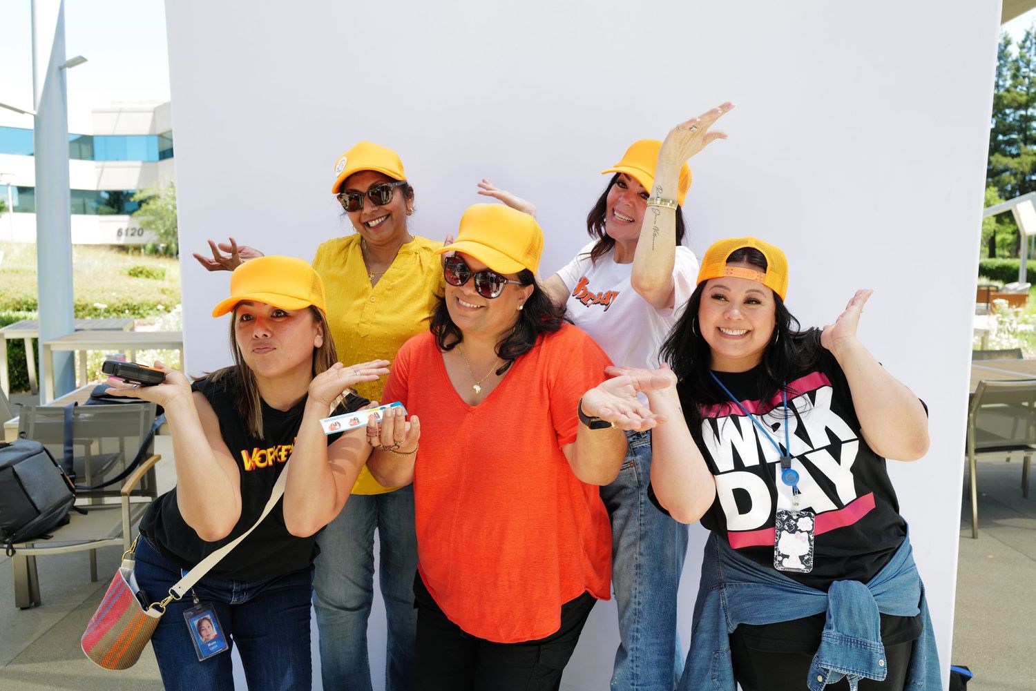 Five women in yellow caps pose playfully at a Workday corporate event outdoors on a sunny day.
