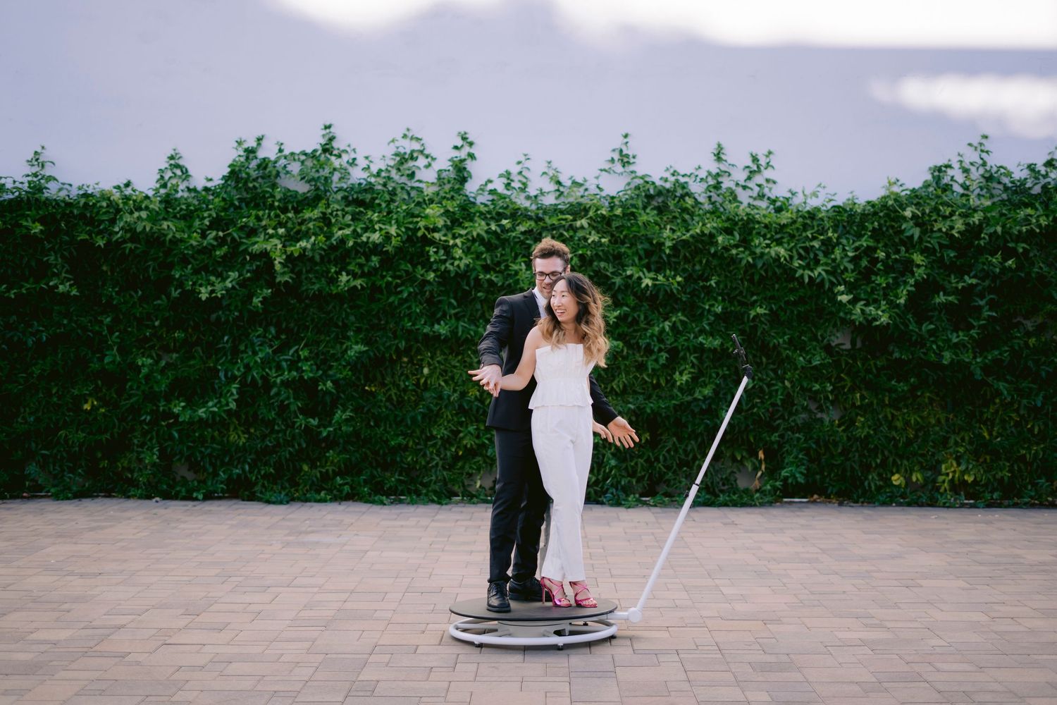 Bride and groom standing on a circular platform outdoors, posing in front of a lush green hedge wall.